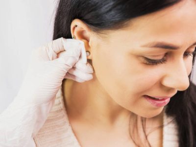 Side view photo of a beautiful brunette woman preparing to a piercing procedure and doctor in sterile gloves rubbing the ear with alcohol. High quality photo