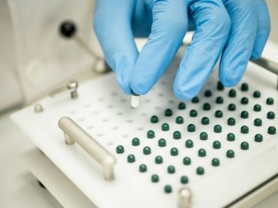High angle of crop anonymous scientist in blue gloves putting capsule into filling machine during process of personalized medications preparation